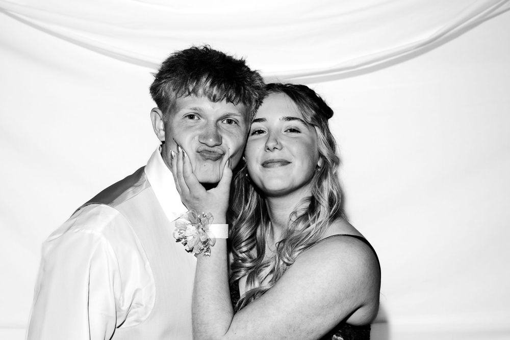 Black and white photo of a young couple smiling together at a photo booth against a white backdrop.
