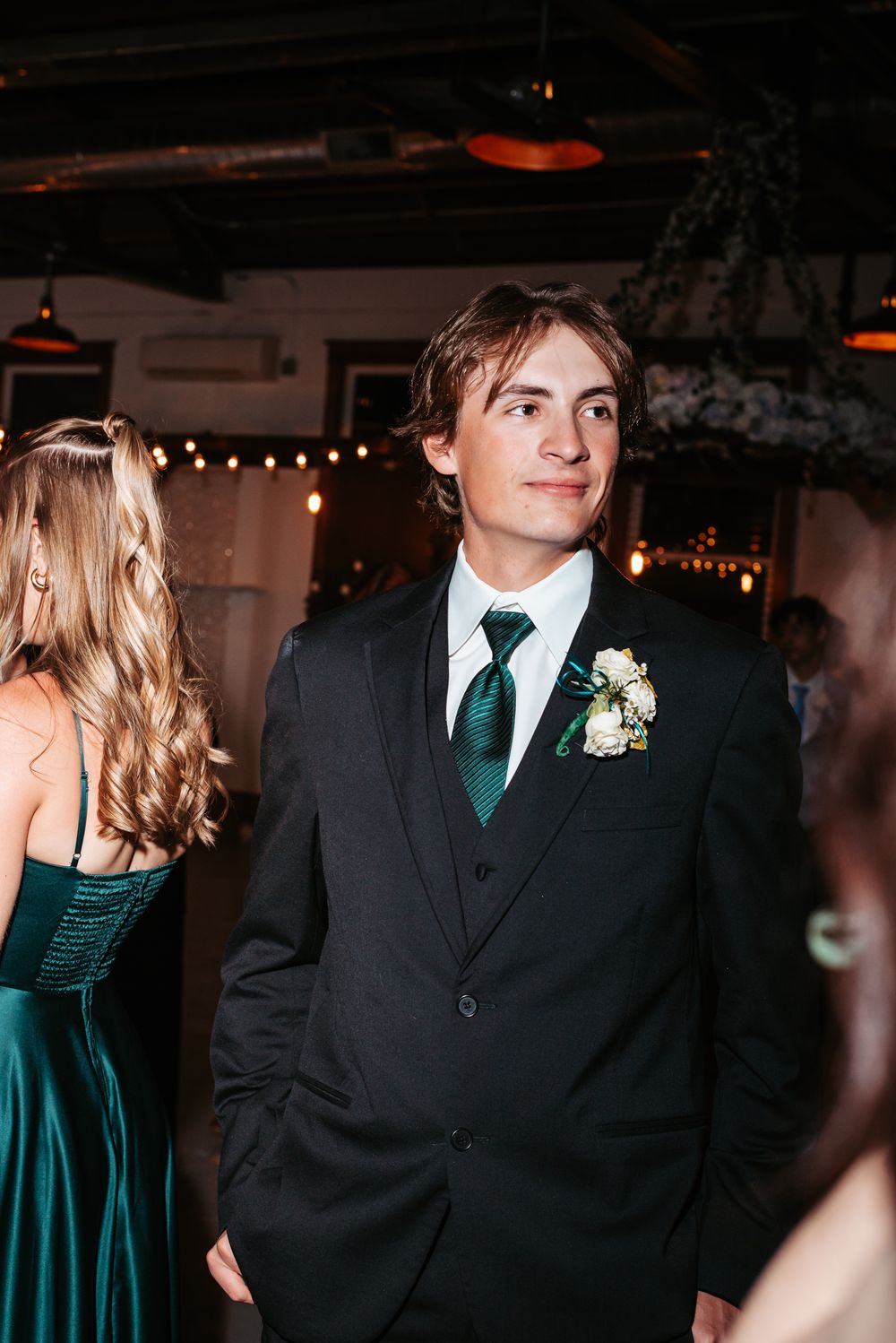 Guest in dark suit with emerald green tie and boutonniere at formal event with string lights and celebration decor.