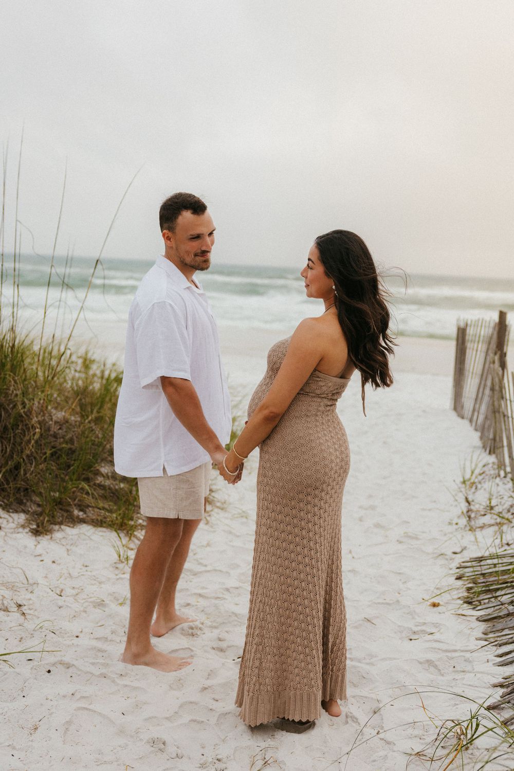 Expecting couple holds hands while standing on sandy beach path with ocean waves behind them.
