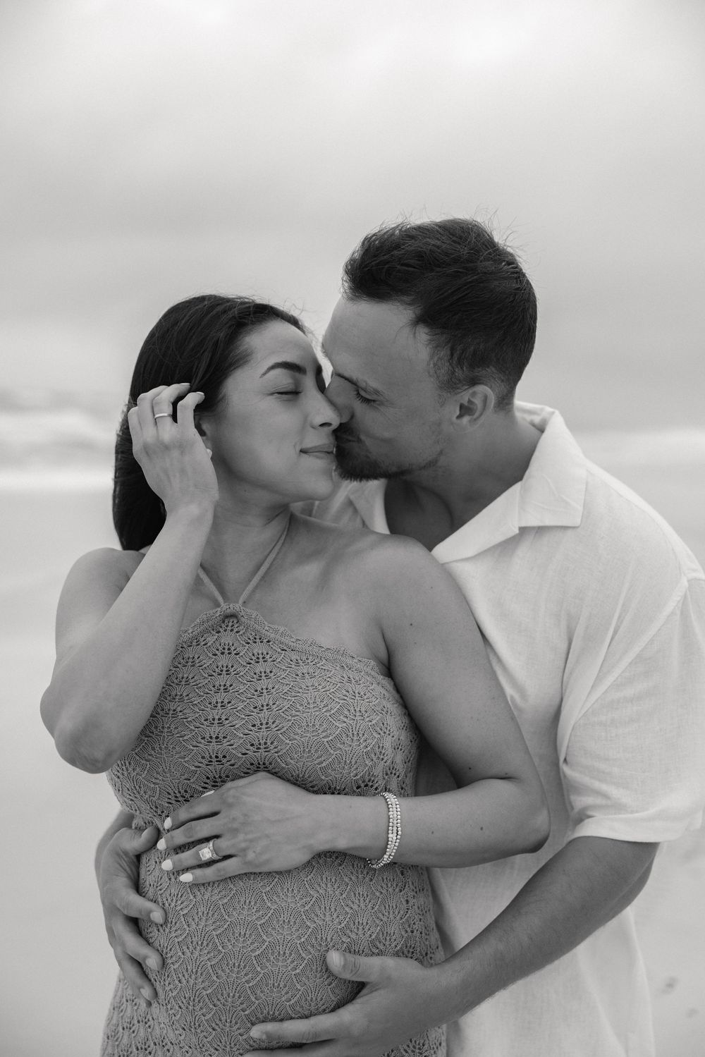 Intimate beach maternity photo of couple embracing in black and white against cloudy sky.