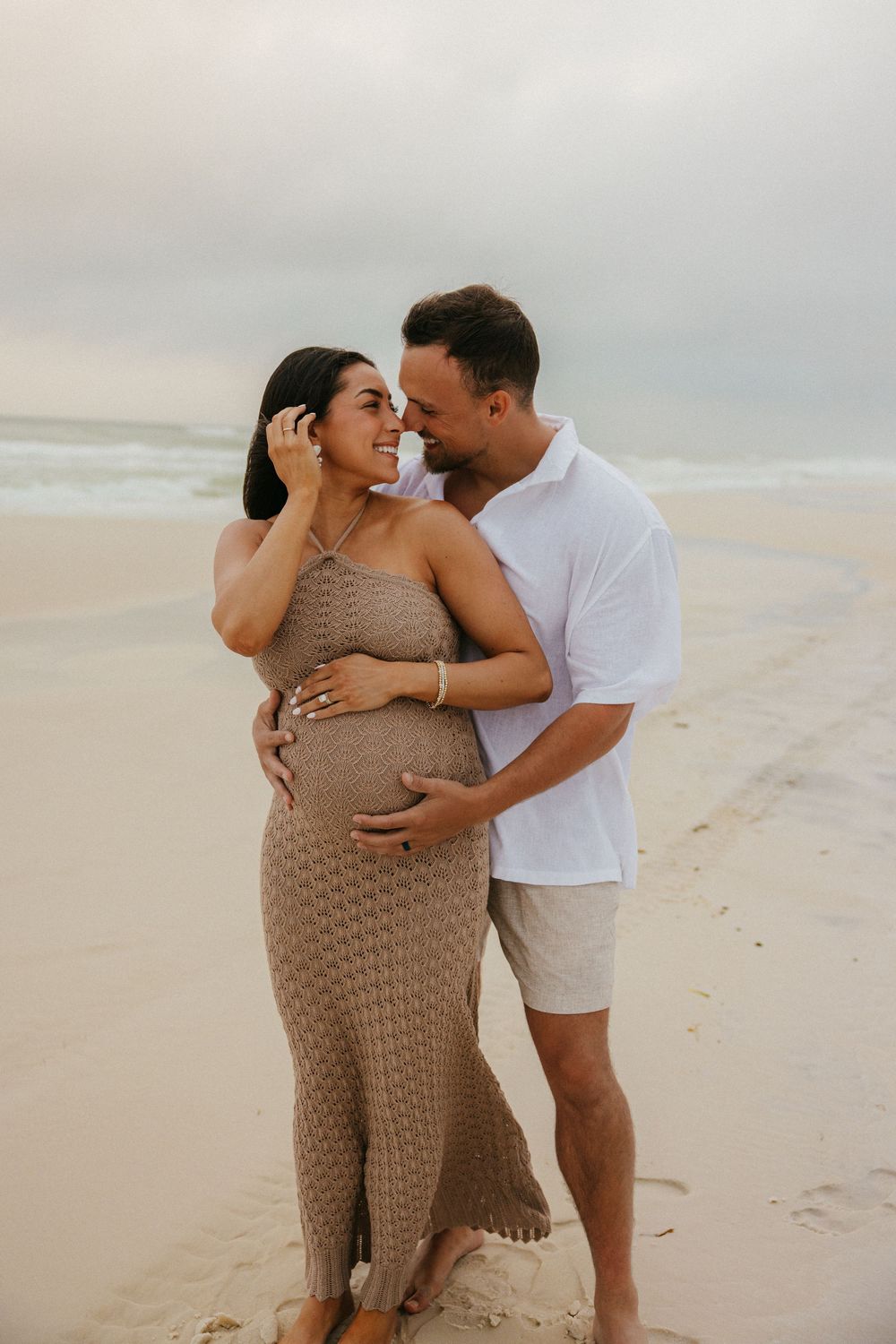 A couple shares an intimate moment on the beach during a maternity photoshoot at sunset.