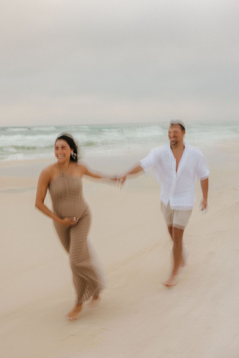 A couple runs playfully along a misty beach shore, with the person in beige and person in white holding hands.