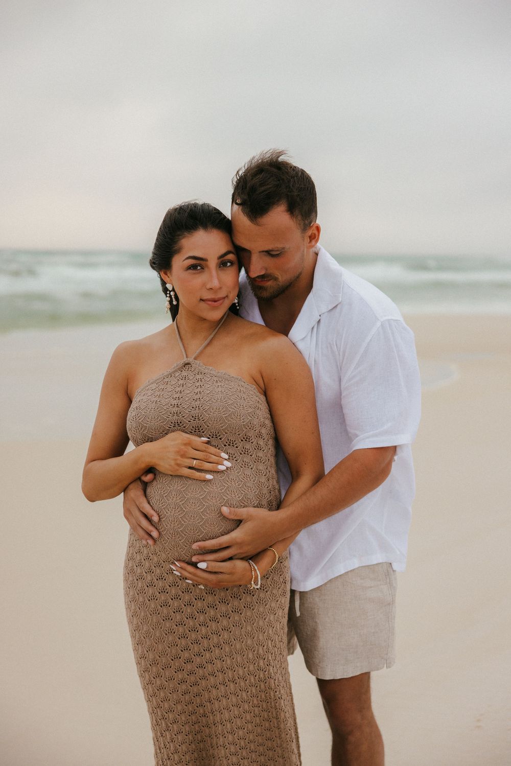 Intimate beachside maternity portrait with couple in white shirt and tan knit dress against ocean backdrop.