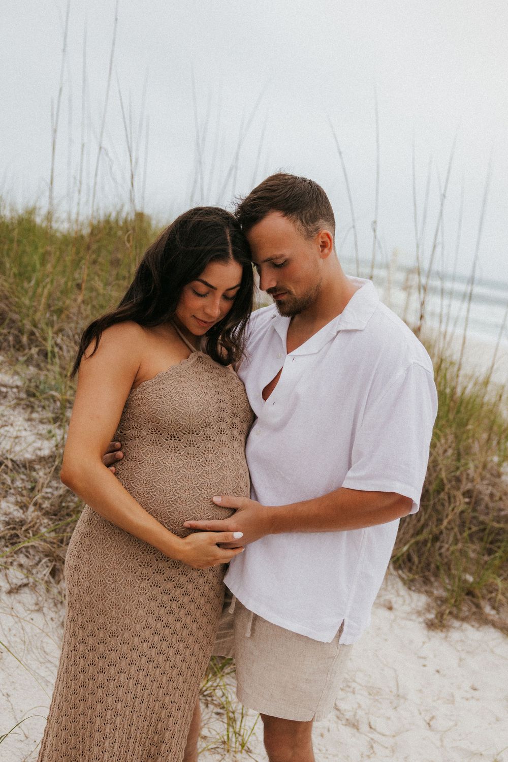 Romantic maternity photo of couple embracing on beach with tall grass and ocean in background.