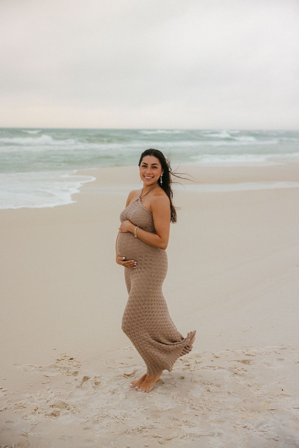 Expectant mother in flowing beige dress smiles while standing barefoot on sandy beach.
