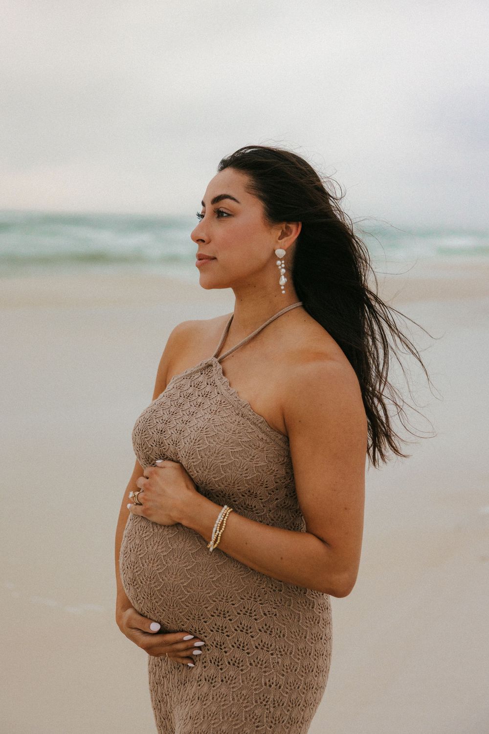 Side profile of an expectant mother in a tan knit dress standing on a beach with wind-swept hair.