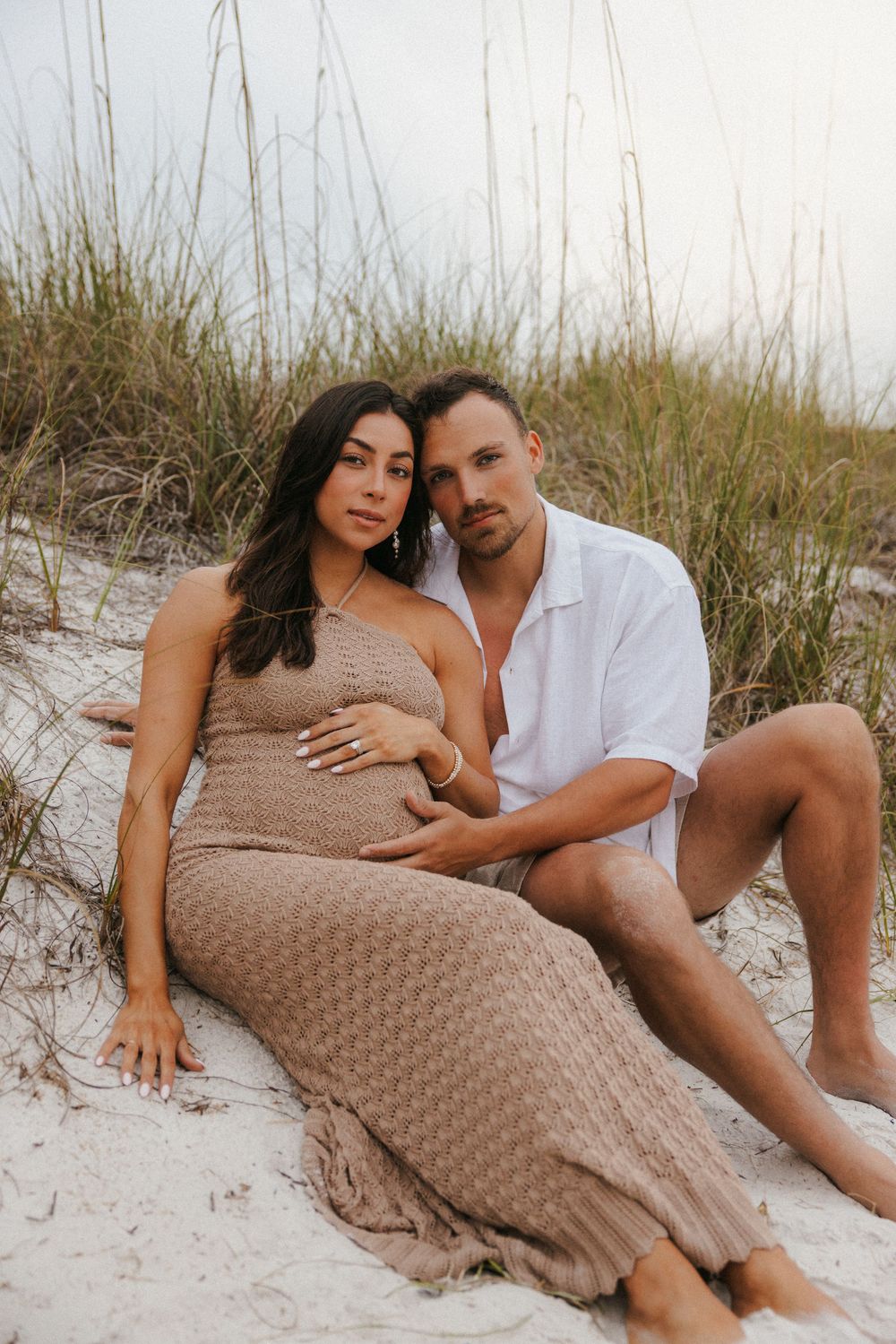 A romantic maternity photo session on a sandy beach with tall grass, featuring a couple in casual beachwear.