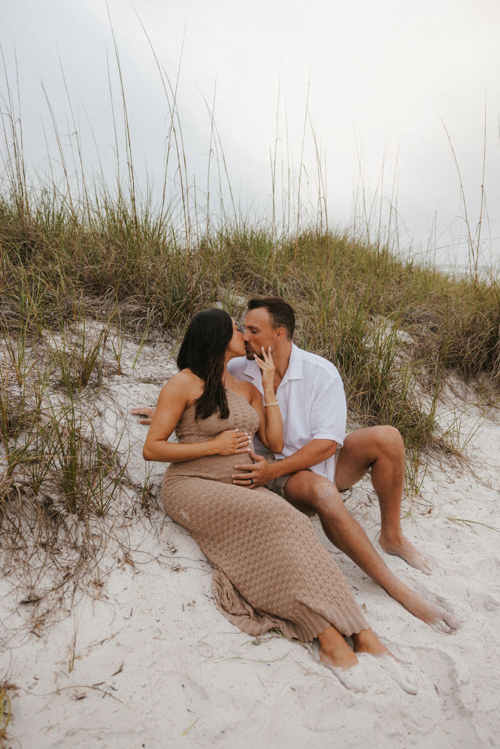 Romantic maternity photo of a couple sitting together in coastal sand dunes.