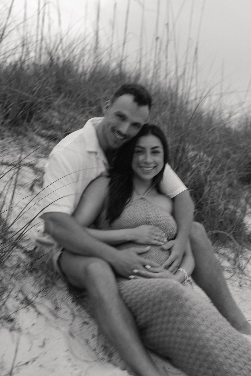 Black and white maternity photo of a couple sitting together on a sand dune.