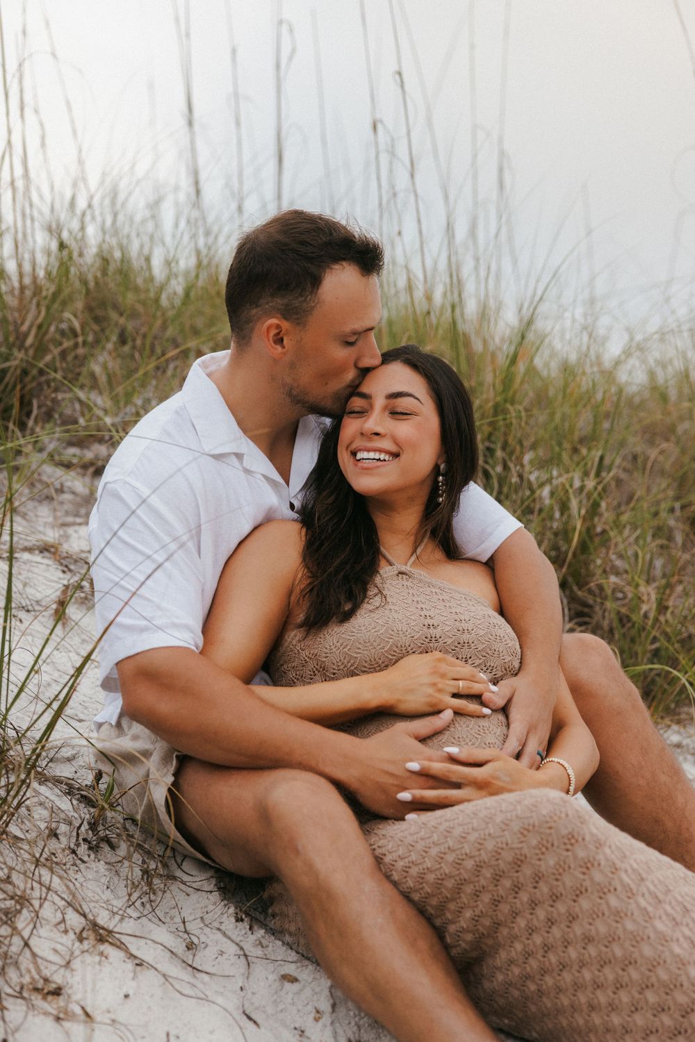Couple sharing intimate moment on sandy dunes during sunset maternity photoshoot.