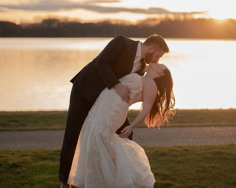 Romantic couple in wedding attire share a passionate dip kiss at sunset by a serene lakeside.