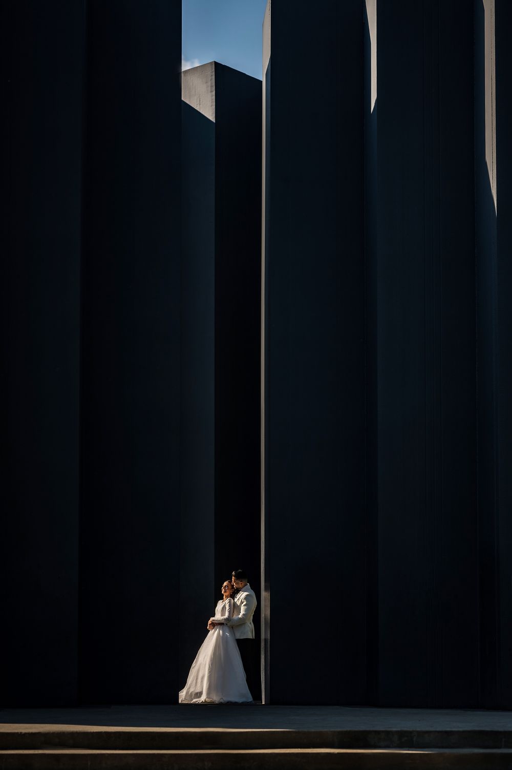 A couple in wedding attire stands in dramatic lighting between tall dark architectural columns creating a striking silhouette.