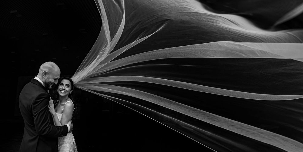 Dramatic black and white wedding photo with flowing veil creating ethereal streaks of light against dark background.