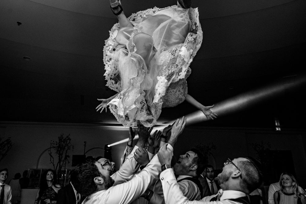 A crowd lifts a bride celebrating at a wedding reception as her dress billows dramatically in black and white.