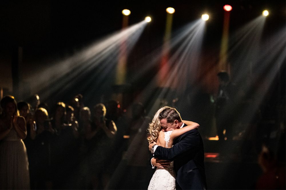 Two people share an intimate dance beneath dramatic spotlights on a dark dance floor surrounded by silhouetted guests.