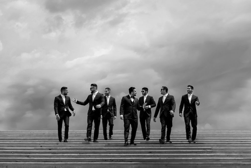 A group of groomsmen in dark suits walk together along stairs under dramatic cloudy skies in black and white.