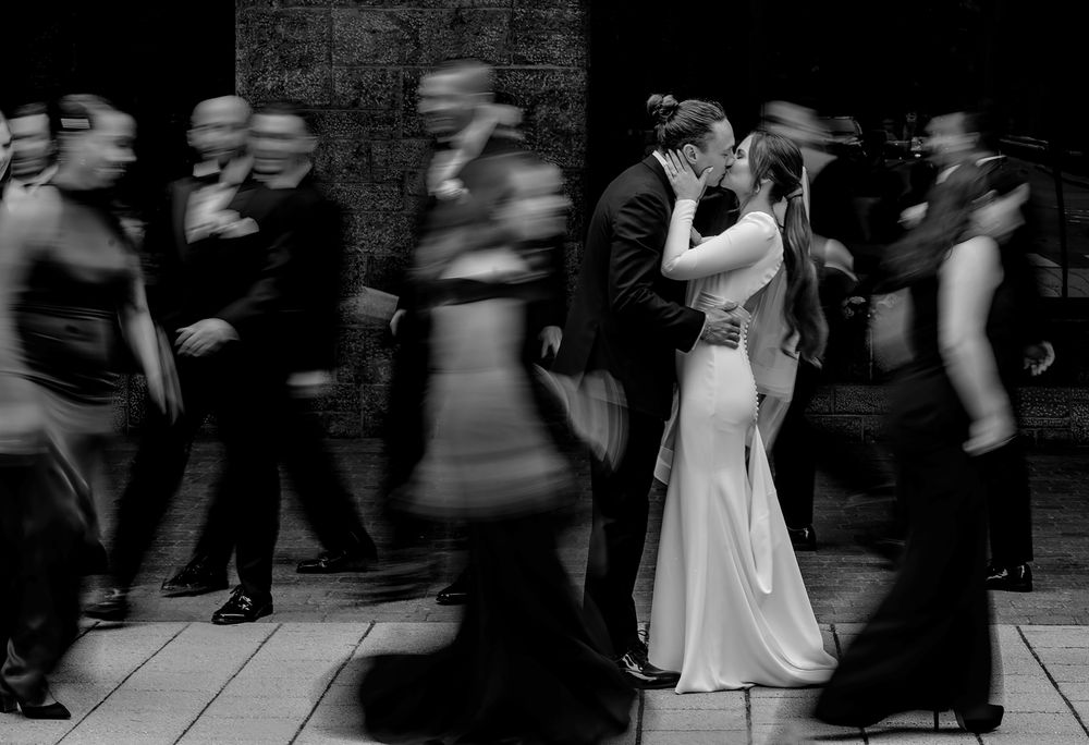 A couple shares a romantic kiss while blurred figures move around them in black and white.
