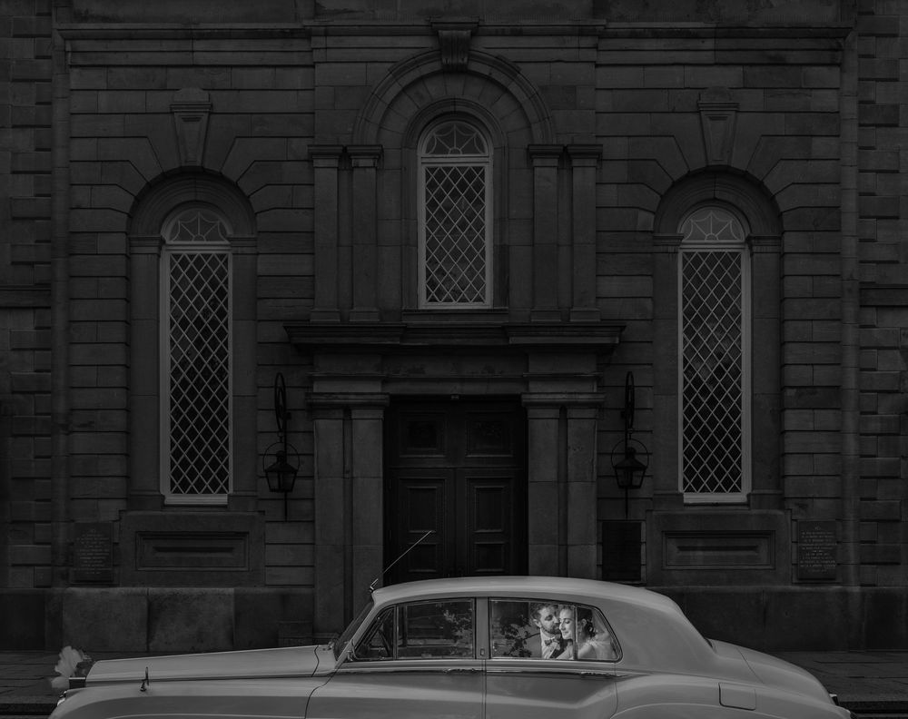 A classic car parked in front of a gothic stone church building with arched windows and ornate doorway.