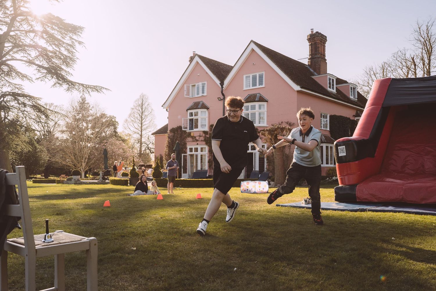 Children play and jump on a bouncy castle in a sunny backyard with a pink house in the background.