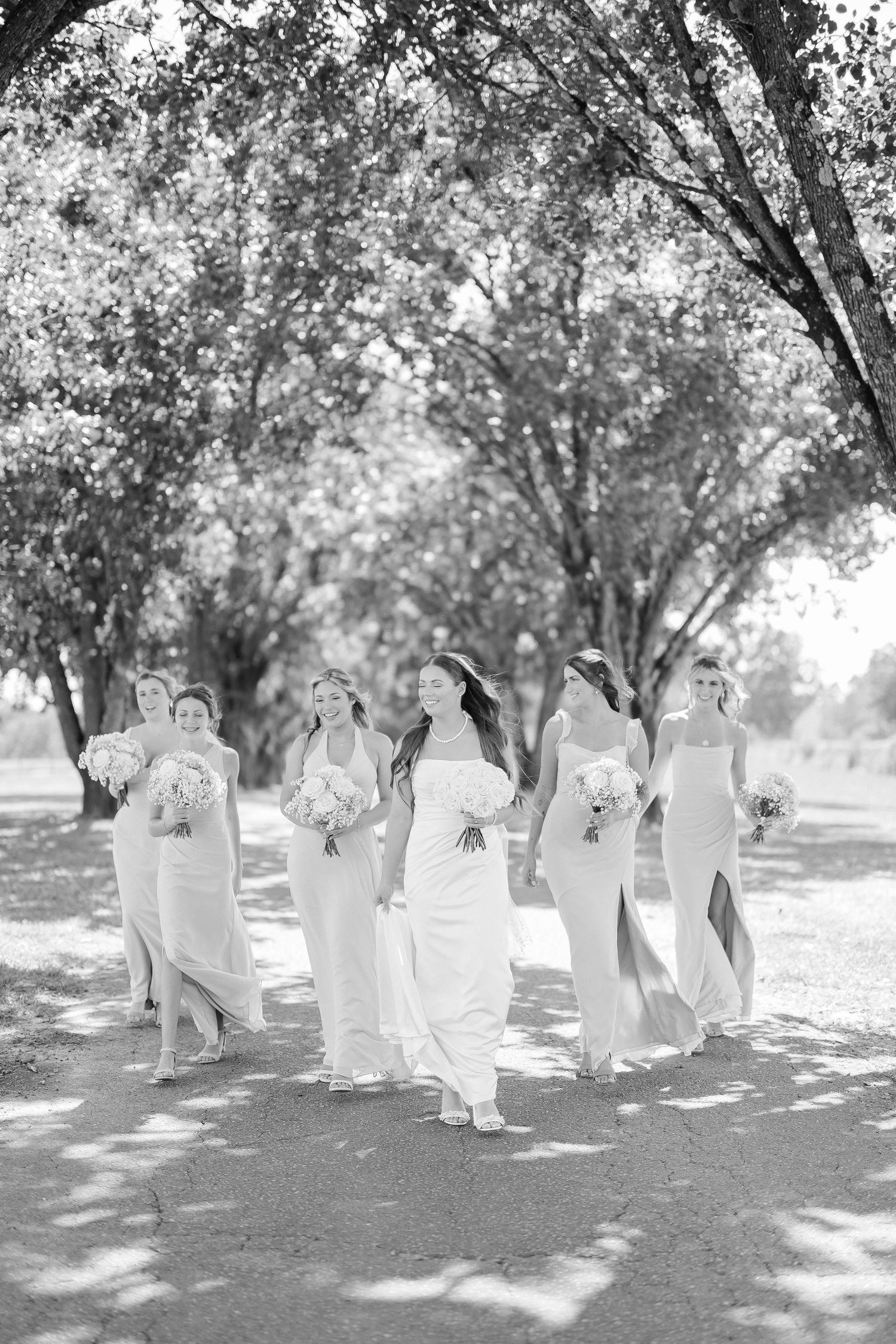 Black and white photo of a bride and bridesmaids in white gowns walking outdoors under lush trees holding bouquets.