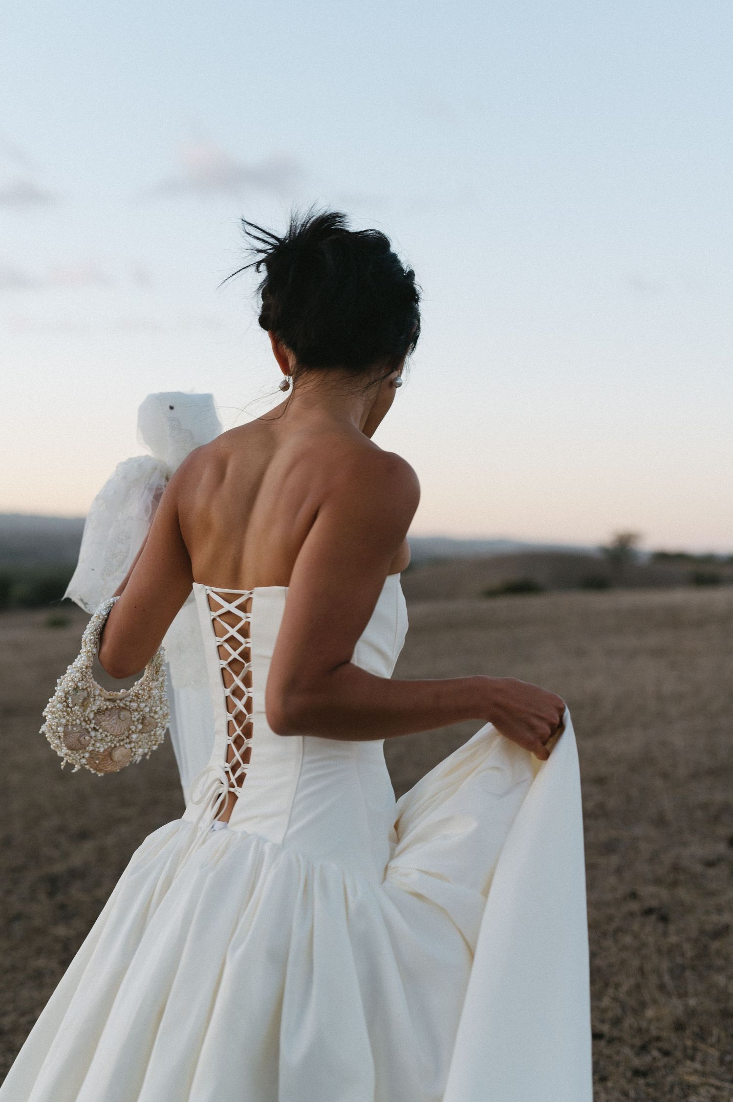 A bride in a white corseted wedding dress with lace-up back detail stands in a field at dusk.