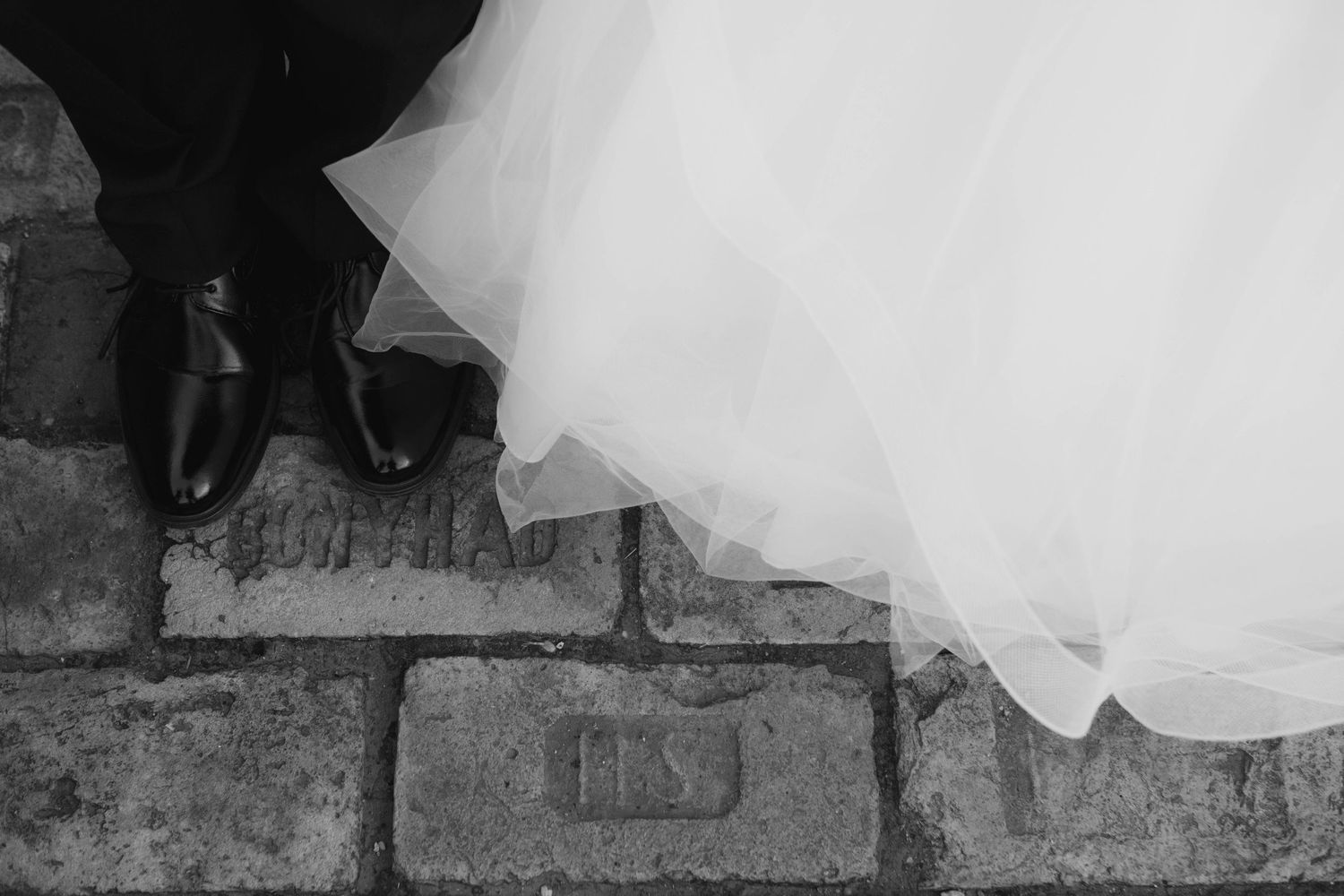 Black and white detail of feet on stone steps with flowing white wedding dress fabric trailing down.
