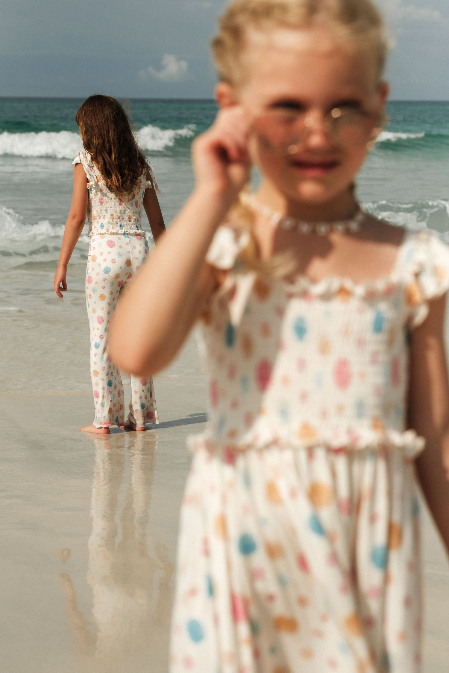 Children in floral dresses playing on a sunny beach with waves in the background.