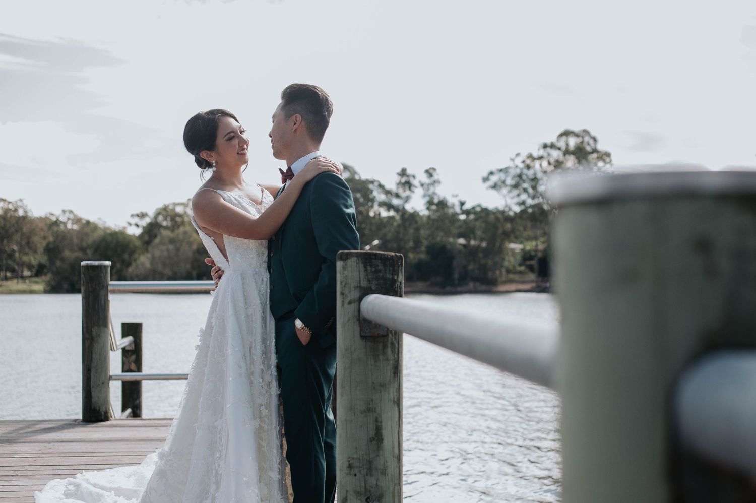 A romantic newlywed couple embraces on a wooden dock overlooking a serene waterfront on their wedding day.