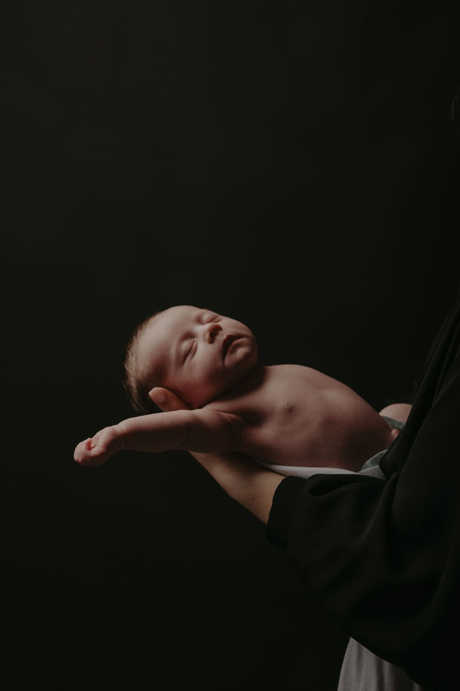 A newborn baby sleeping peacefully against a dark background in a minimalist artistic portrait.