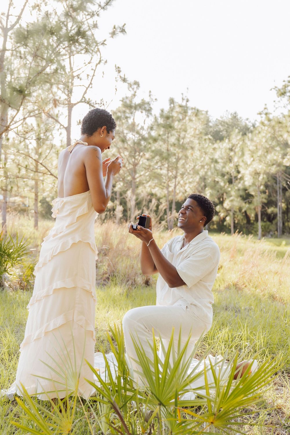 Romantic proposal scene in a sun-drenched field with palm fronds, featuring two people in elegant white outfits.