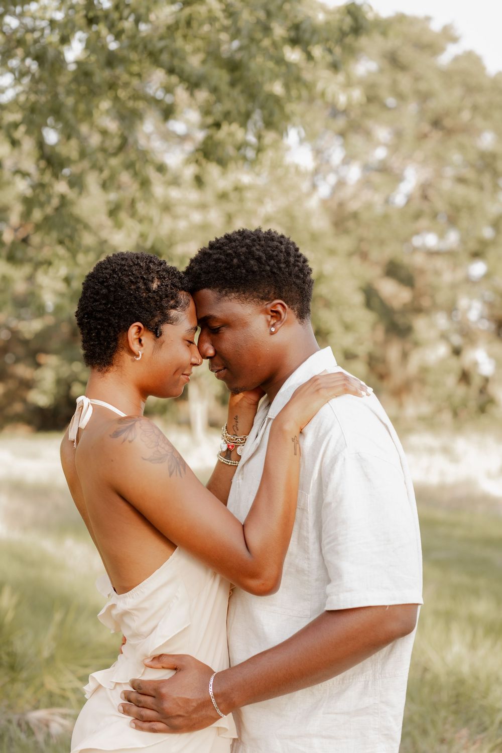 A romantic outdoor portrait of a couple embracing in soft sunlight with trees in the background.