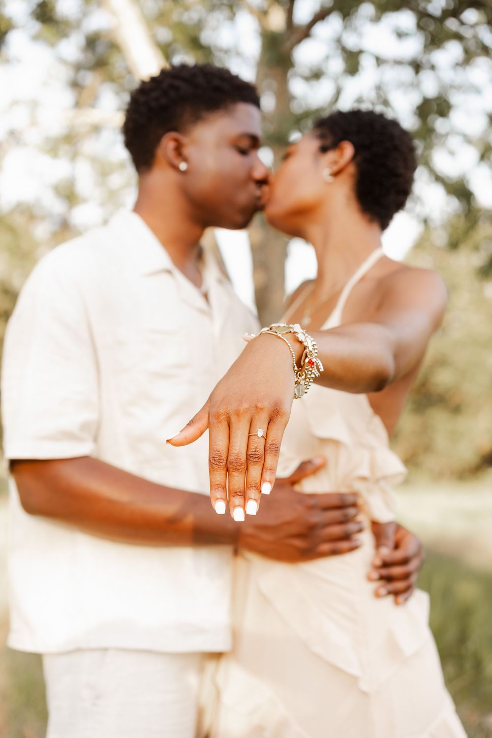 A romantic couple in white outfits embrace outdoors in a dreamy garden setting showing off an engagement ring.