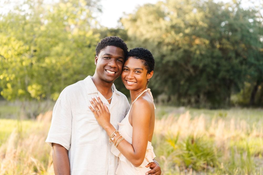 A happy couple embraces and smiles together in a sunlit field with golden grass and trees in the background.