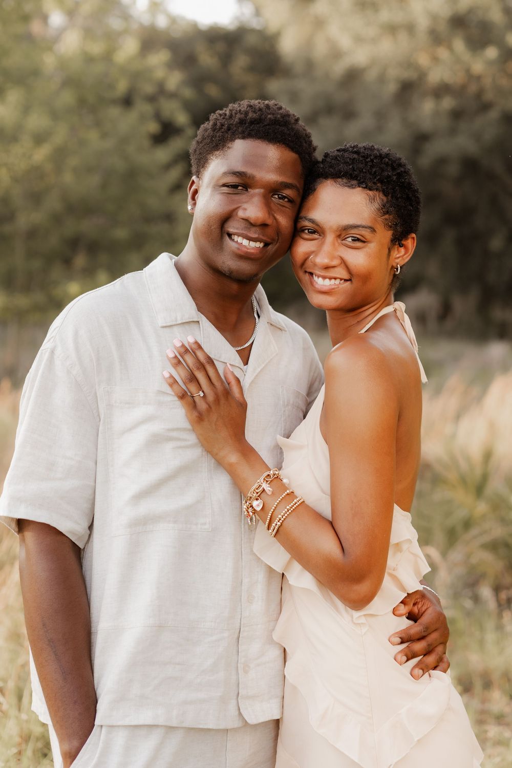 A couple in white outfits embrace outdoors during a romantic photoshoot with natural lighting and greenery in background.