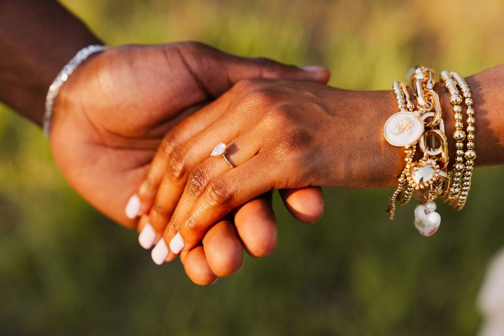 Two hands clasped together romantically outdoors, with one wrist wearing gold charm bracelets and the other wearing a diamond ring.