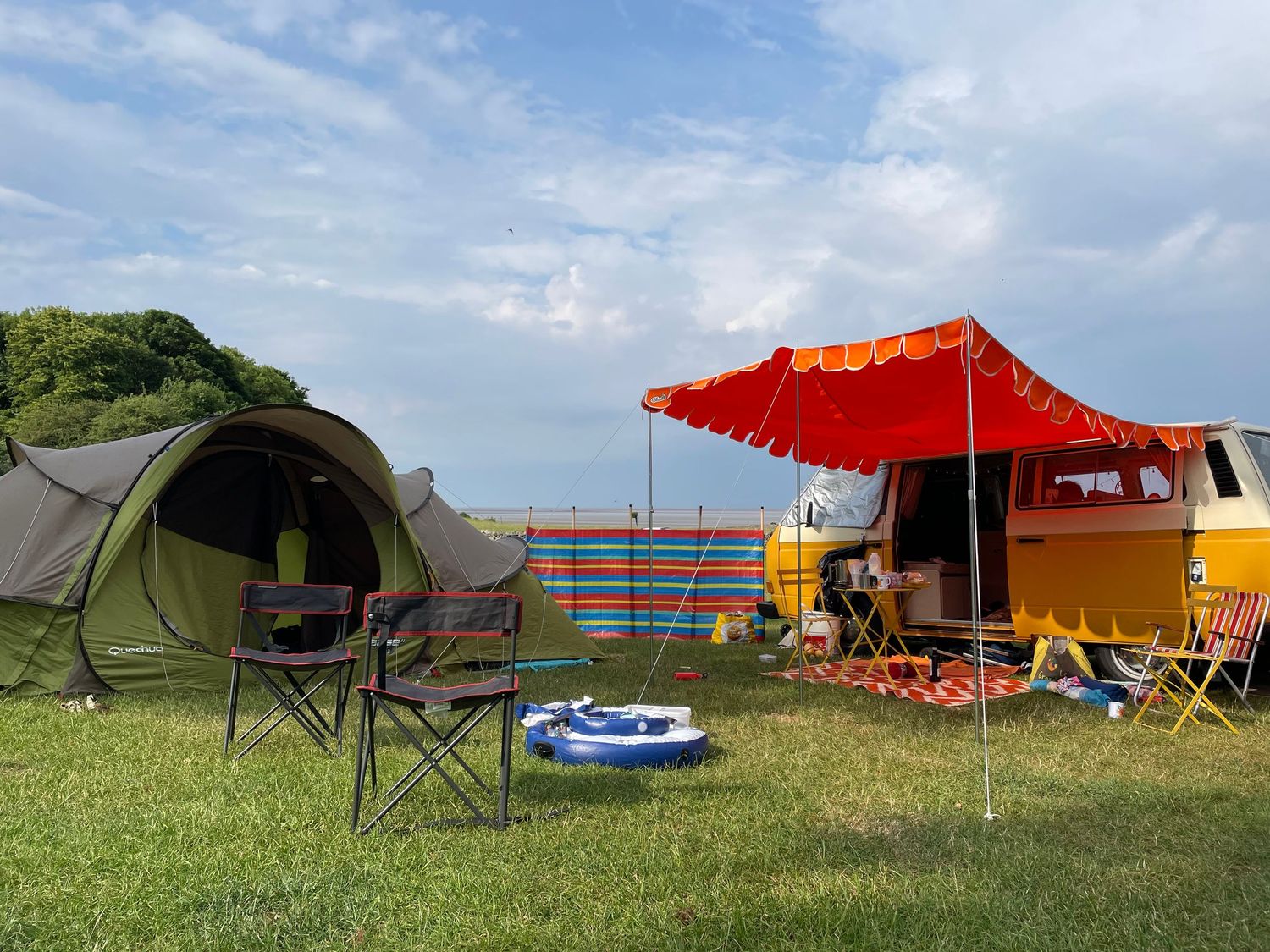 Campsite setup with green tent and orange awning van parked on grassy hill overlooking ocean view on sunny day.