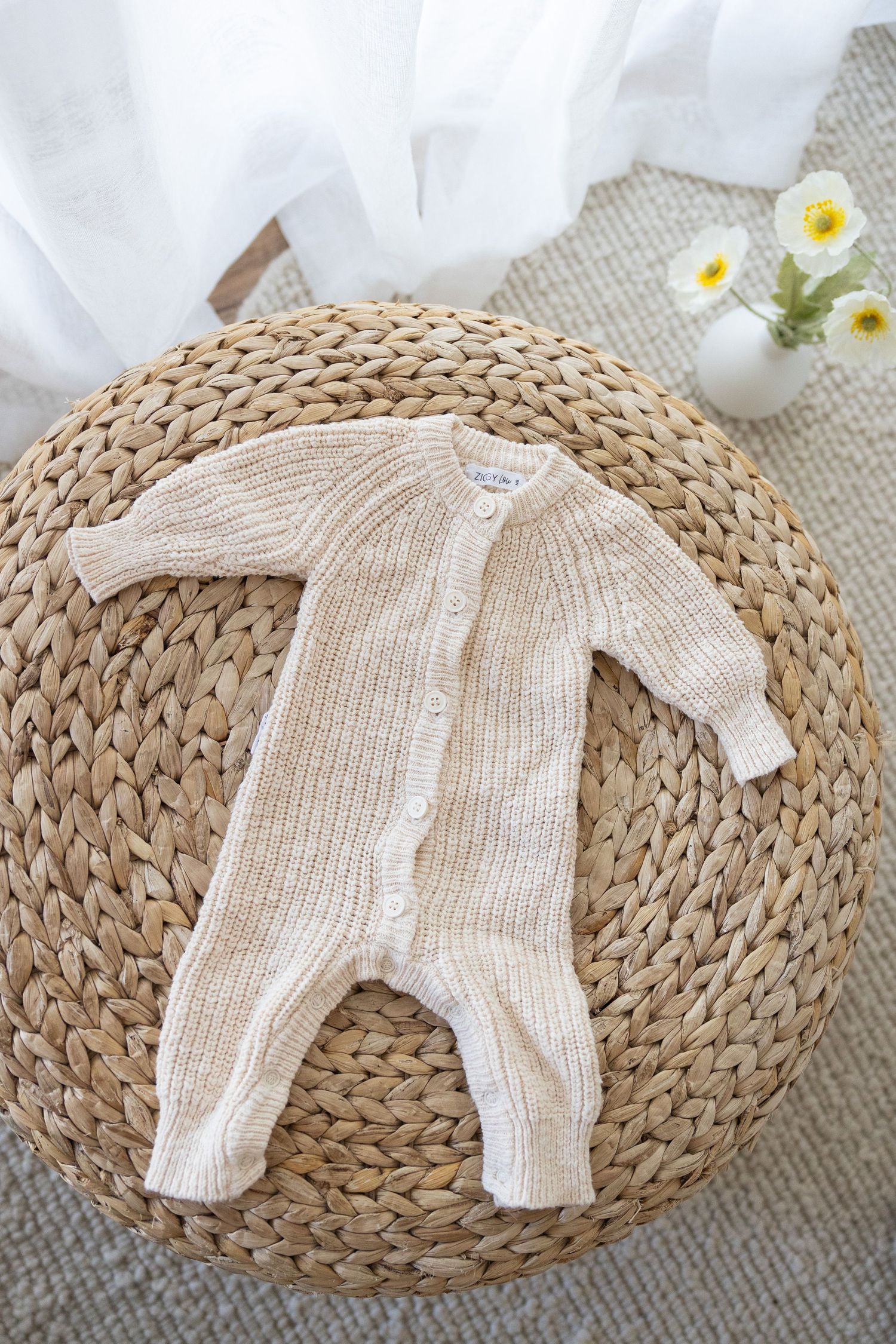 Knitted cream-colored baby romper displayed on a round woven rattan mat with white flowers in the background.