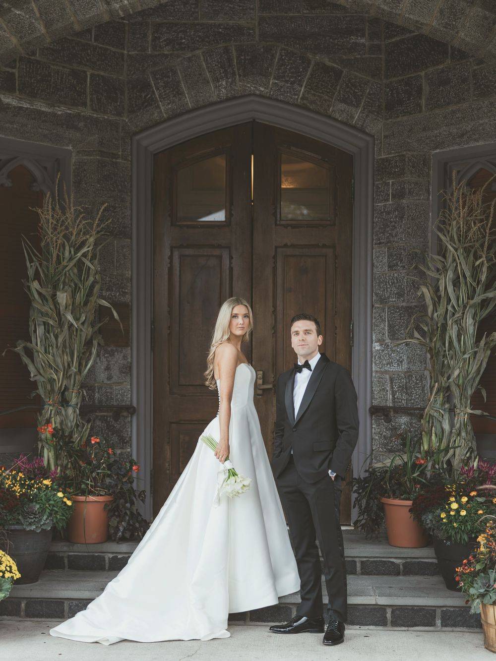 A wedding couple poses outside a stone building entrance decorated with potted plants and floral arrangements.