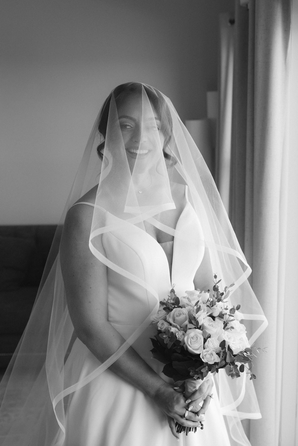 Black and white portrait of a smiling bride wearing a flowing veil and holding a bouquet of roses.