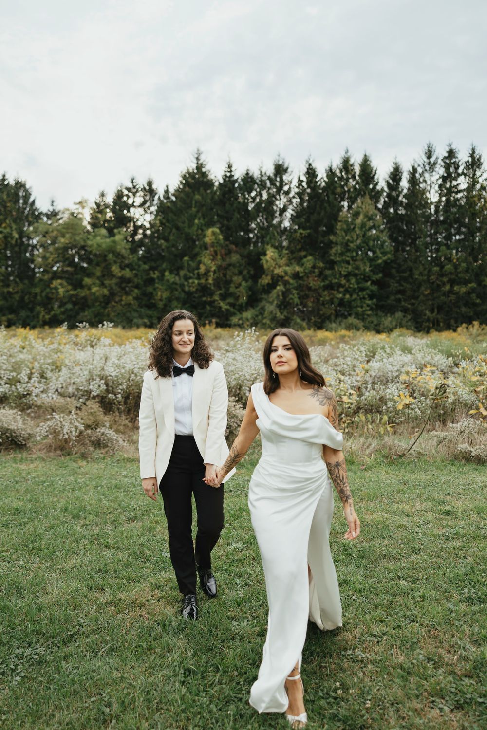 A couple in wedding attire walks hand in hand through a wildflower meadow with evergreen trees in the background.