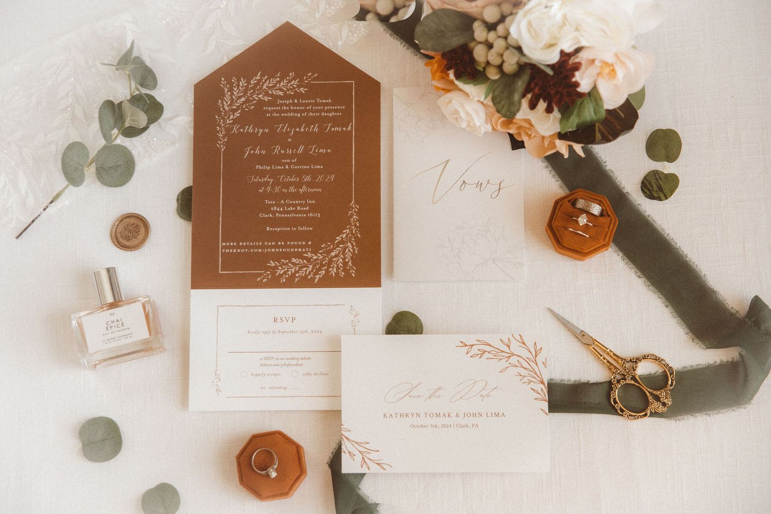 An image of a flat lay of wedding details, including an invitation set, vow book, rings in a ring box, ribbon, and perfume.