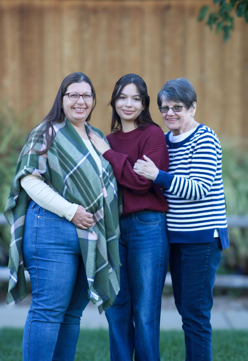 Three generations pose casually outdoors wearing sweaters and jeans against a wooden fence background.