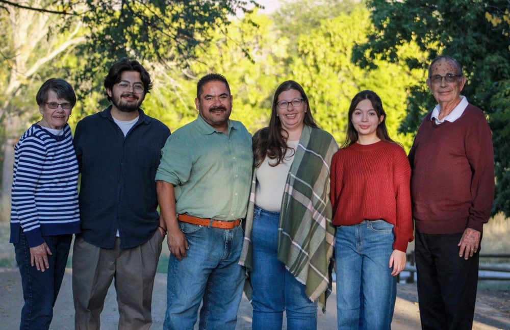 Group of six people standing together outdoors in casual attire under trees.