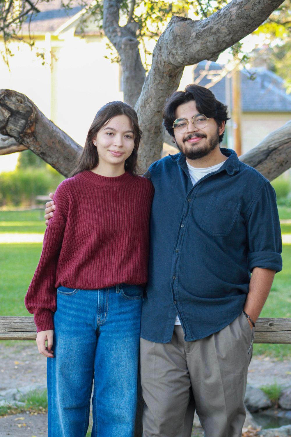 A couple poses together by a large tree trunk in outdoor garden setting.