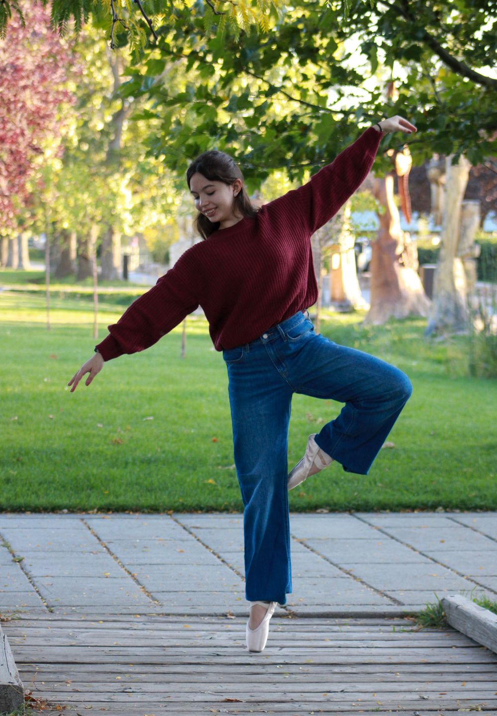 Dancer in red sweater and jeans strikes graceful pose on wooden boardwalk.