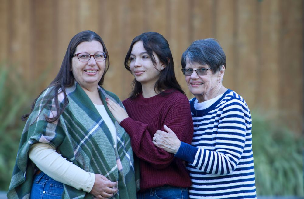 Small family group smiles together outdoors wearing casual clothing against wooden backdrop.