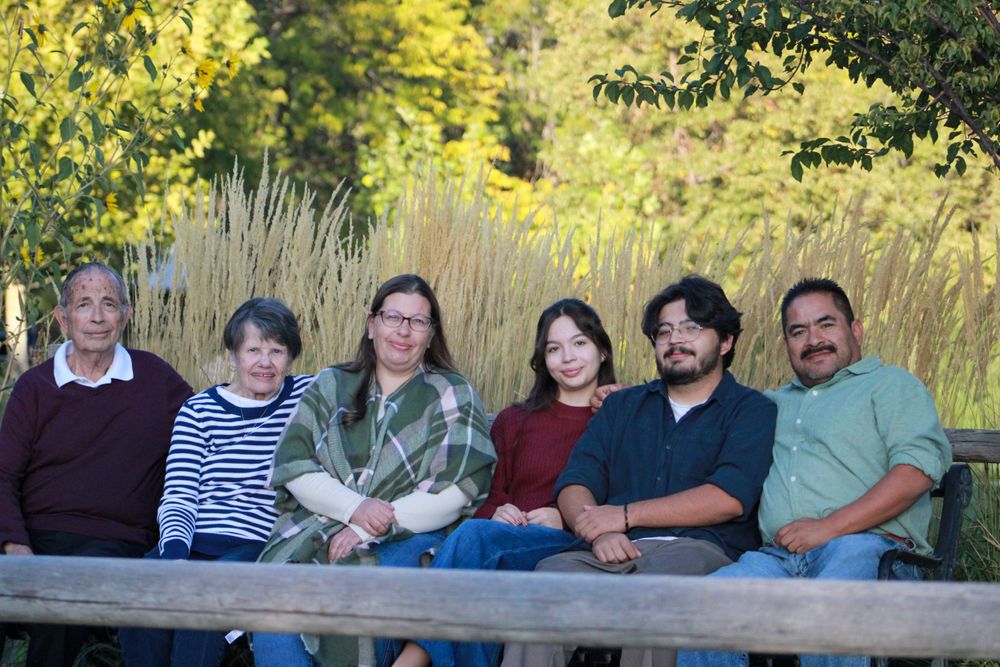 Group of six people sit casually on wooden bench with autumn foliage in background.