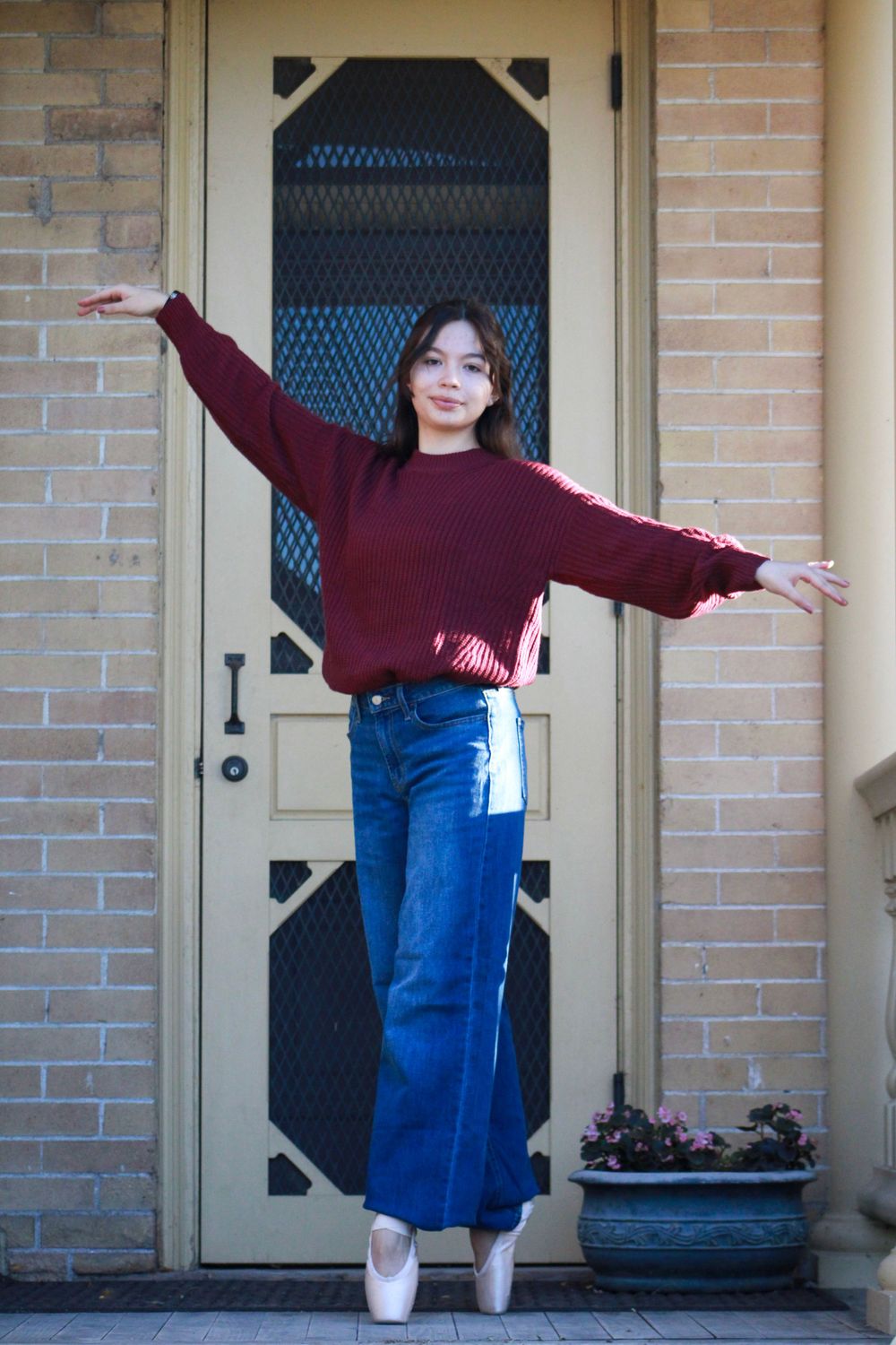Dancer in burgundy sweater and jeans strikes graceful pose in ballet shoes on front porch.