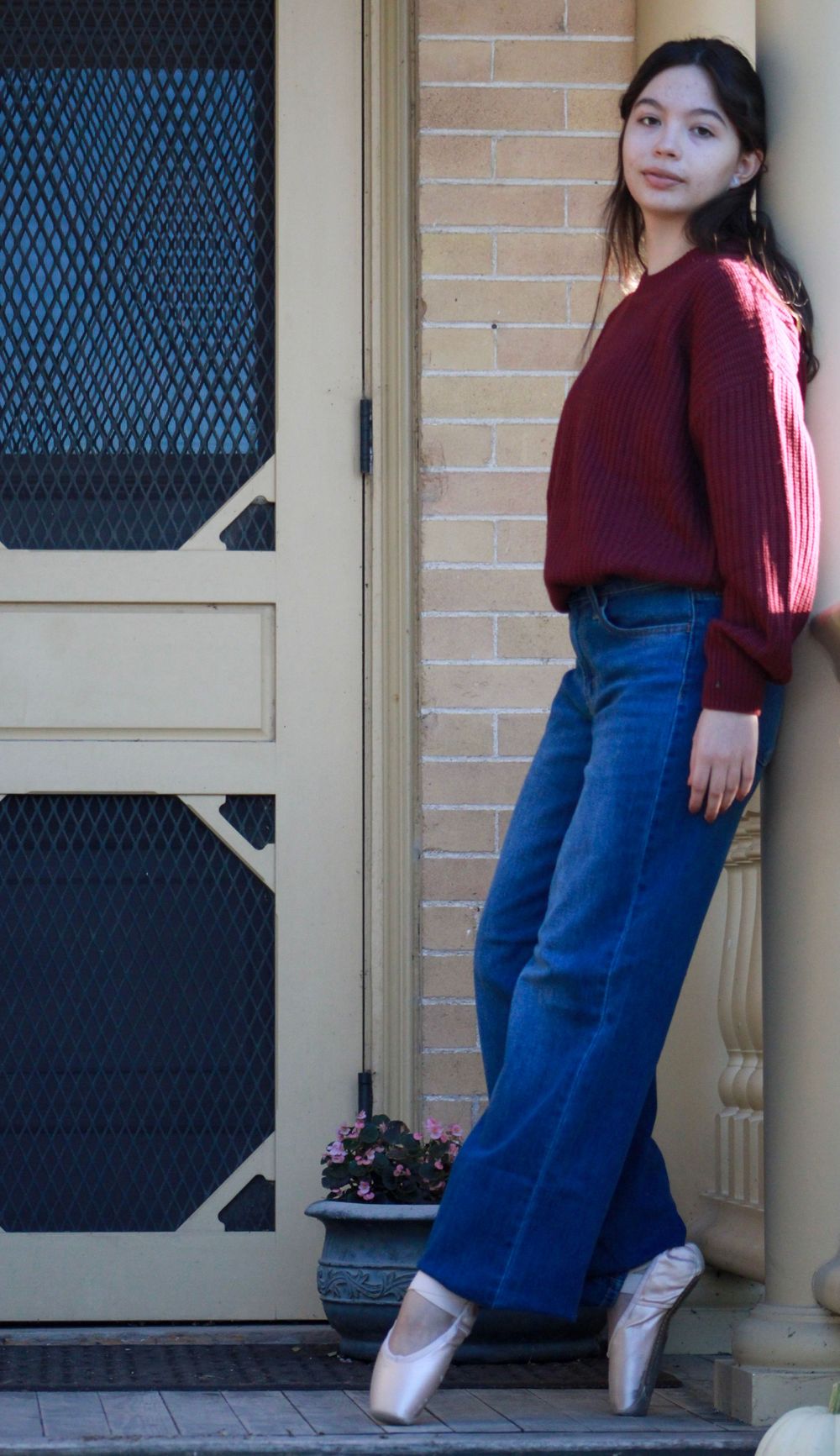 Ballet dancer in casual attire poses en pointe against brick wall and screen door.