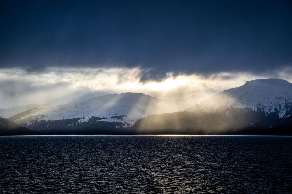 Dramatic sunbeams pierce through storm clouds over a mountainous lake landscape.